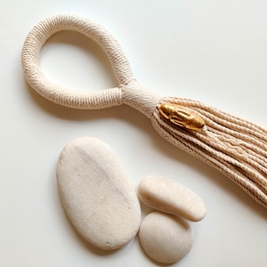 May include: A beige macrame wall hanging with a gold bead accent. The hanging is made of a thick, twisted cord and has a tassel at the bottom. The hanging is displayed against a white background with three white stones.