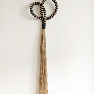 May include: A decorative wall hanging featuring a looped design with black and tan rope. The loops are connected by a black band, with a long, natural-colored tassel hanging below. This piece adds a touch of bohemian style to any space.