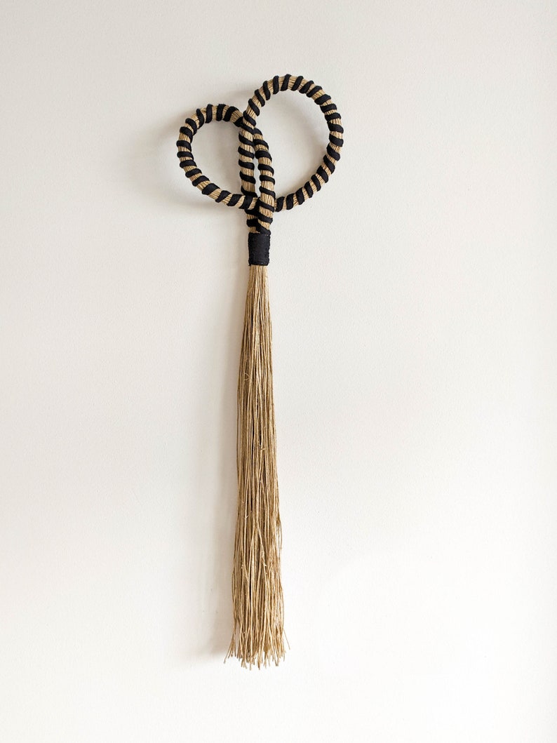 May include: A decorative wall hanging featuring a looped design with black and tan rope. The loops are connected by a black band, with a long, natural-colored tassel hanging below. This piece adds a touch of bohemian style to any space.