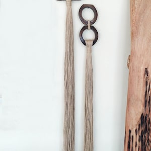May include: Two wooden wall hangings with natural jute tassels. The larger hanging is a triangular shape with a circular cutout. The smaller hanging is two connected circles.