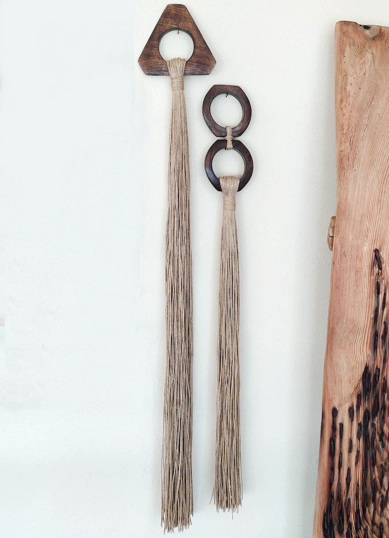 May include: Two wooden wall hangings with natural jute tassels. The larger hanging is a triangular shape with a circular cutout. The smaller hanging is two connected circles.