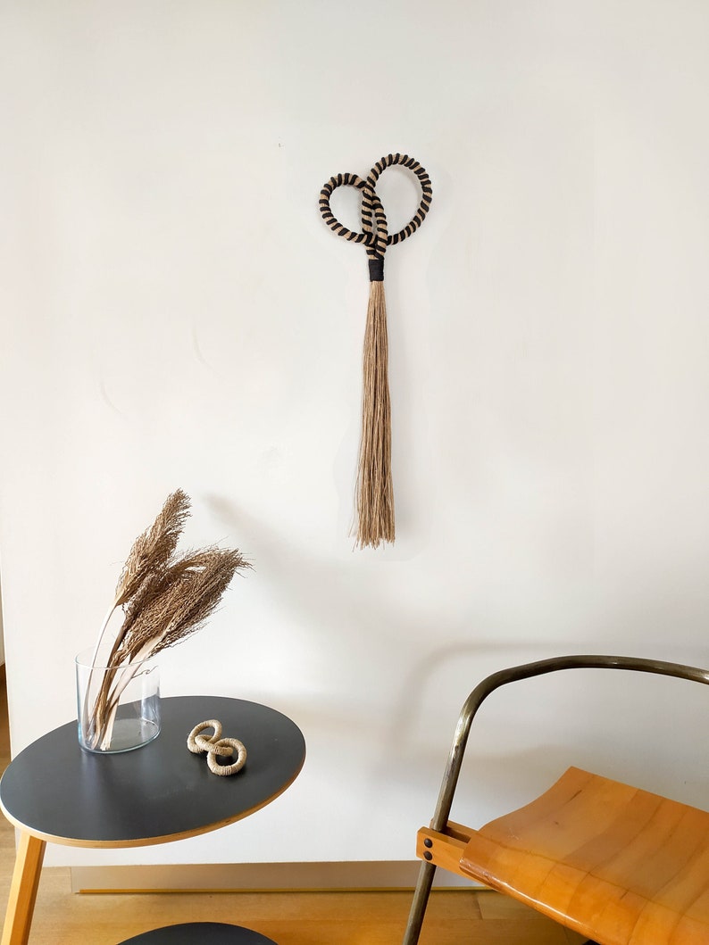 May include: A woven wall hanging with a heart-shaped top and a long, fringed bottom. The top is black and tan rope, and the fringe is a natural tan color. The wall hanging is displayed on a white wall.