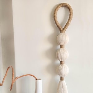 Könnte beinhalten: Handgefertigte Wanddekoration mit einer Schlaufe aus Naturjute, drei großen, cremefarbenen Garnkugeln und einer Quaste. Die Kugeln sind durch Juteseil getrennt. Dieses dekorative Stück verleiht einen Hauch von Bohème-Stil.
