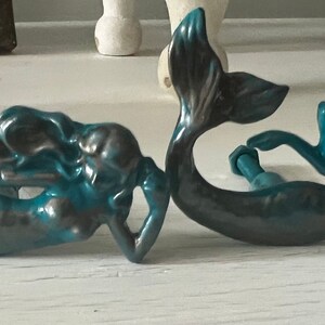 May include: Two teal blue mermaid figurines with a metallic finish. The mermaids are in a curled position with their tails intertwined.