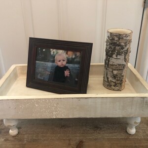 May include: White wooden tray with a distressed finish and turned legs. The tray has a picture frame with a photo of a child and a birch bark cylinder on top.