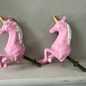 May include: Two pink unicorn-shaped drawer pulls with gold horns and white manes. The pulls are attached to metal screws.
