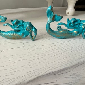 May include: Two turquoise mermaid-shaped cabinet pulls with gold accents. The mermaid figures are in a reclining pose, with their tails curved upwards. The pulls are made of a glossy material and are likely ceramic or porcelain.