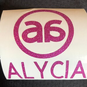 Pode incluir: Adesivo de vinil rosa com glitter com as letras "a6" em um círculo e o nome "ALYCIA" abaixo.