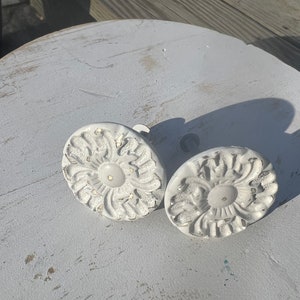 May include: Two white ceramic drawer pulls with a floral design. The pulls are round and have a raised center.