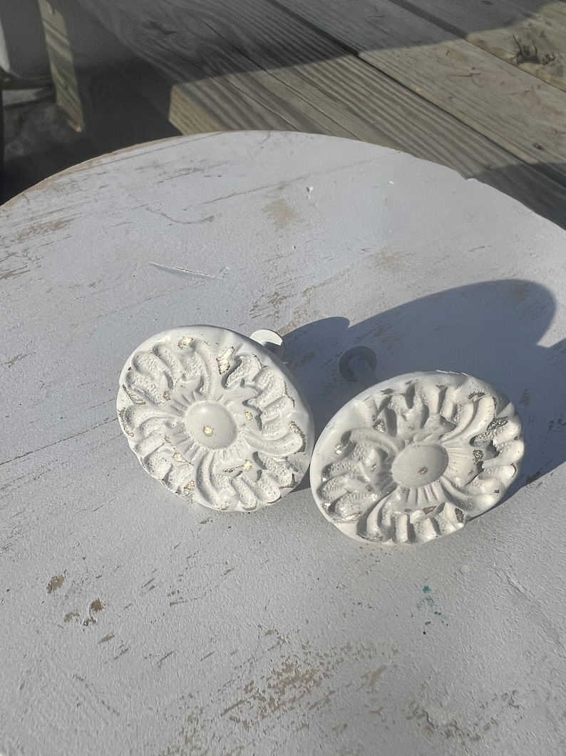 May include: Two white ceramic drawer pulls with a floral design. The pulls are round and have a raised center.