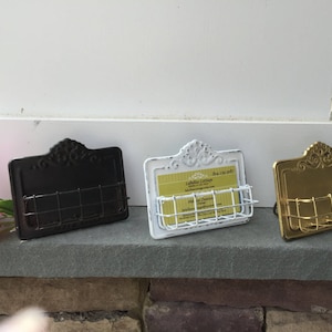 May include: Three metal business card holders with decorative scrollwork designs. The holders are in black, white, and gold.