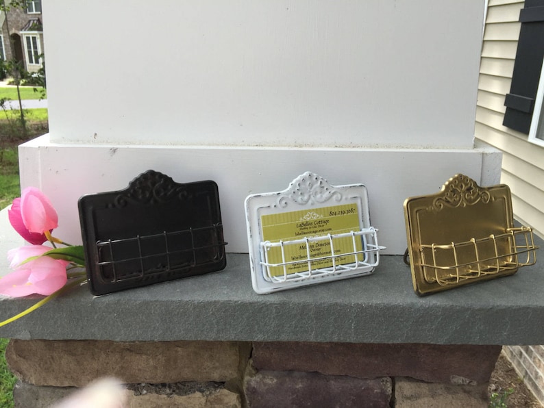May include: Three metal business card holders with decorative scrollwork designs. The holders are in black, white, and gold.