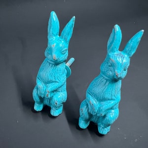 May include: Two turquoise blue ceramic bunny rabbit figurines. The bunnies are sitting upright with their paws crossed. They have long ears and a distressed finish.
