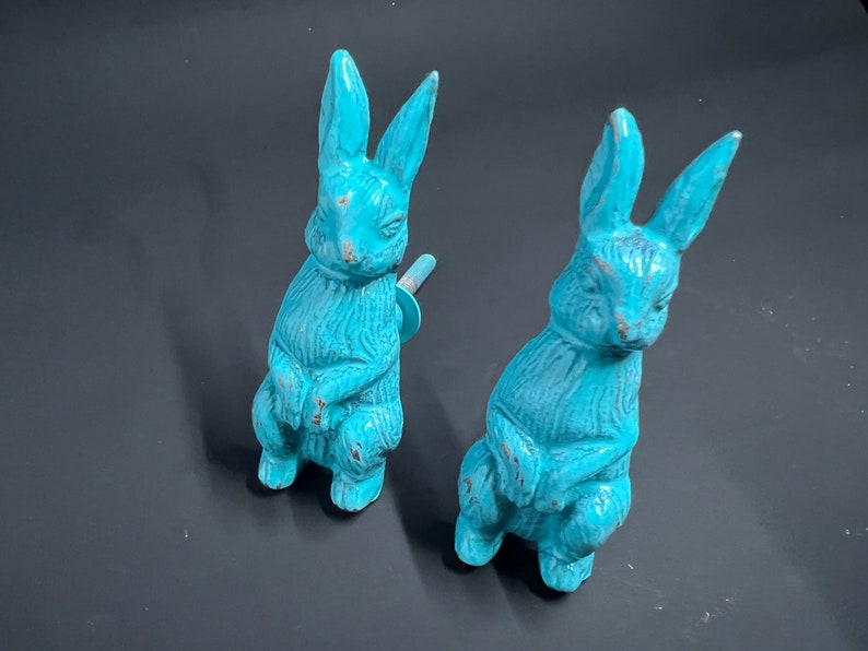 May include: Two turquoise blue ceramic bunny rabbit figurines. The bunnies are sitting upright with their paws crossed. They have long ears and a distressed finish.