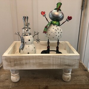 May include: A white wooden tray with four legs, decorated with a white ceramic snowman with a silver hat and a green scarf and a silver metal snowman with a green scarf and red mittens.