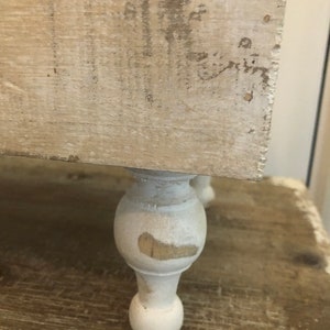 May include: White wooden shelf with three turned legs. The shelf has a distressed white paint finish and is sitting on a wooden surface.