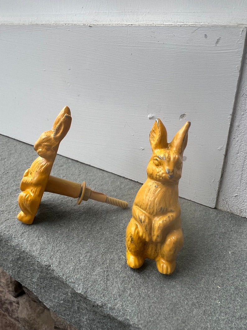 May include: Two yellow ceramic rabbit door knobs. The rabbit on the left is facing the left and the rabbit on the right is facing the right. Both rabbits have their paws crossed in front of them.