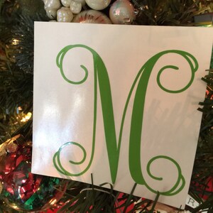 May include: A white square with a green letter "M" in a decorative font. The letter has a swirled design at the top and bottom. The square is displayed on a Christmas tree with ornaments.
