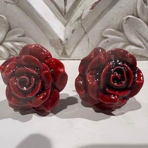 May include: Two red ceramic rose-shaped drawer pulls. The roses have a dark red center and are detailed with petals.