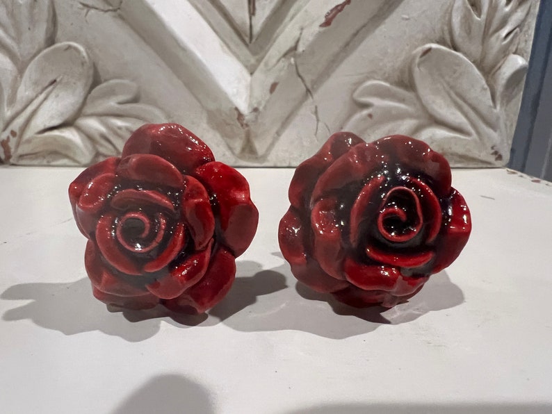 May include: Two red ceramic rose-shaped drawer pulls. The roses have a dark red center and are detailed with petals.
