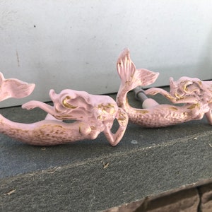 May include: Two pink mermaid-shaped cabinet knobs with gold accents. The mermaids are in a curled position with their tails curled around their bodies.