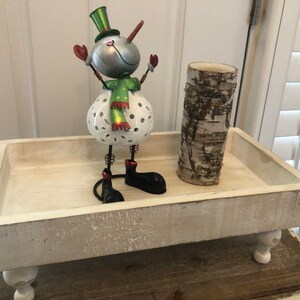 May include: White wooden tray with a snowman figurine and a birch bark candle holder. The tray has four legs and a distressed white finish.