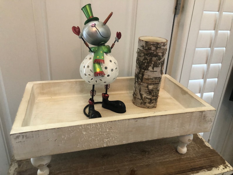 May include: White wooden tray with a snowman figurine and a birch bark candle holder. The tray has four legs and a distressed white finish.