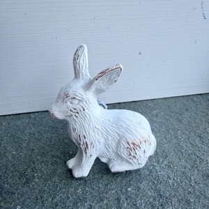 May include: A white rabbit figurine with a textured surface and visible brown distressed detailing on the ears, nose, and fur. The rabbit is depicted in a seated position, showing detailed fur patterns and pointed ears facing upward.