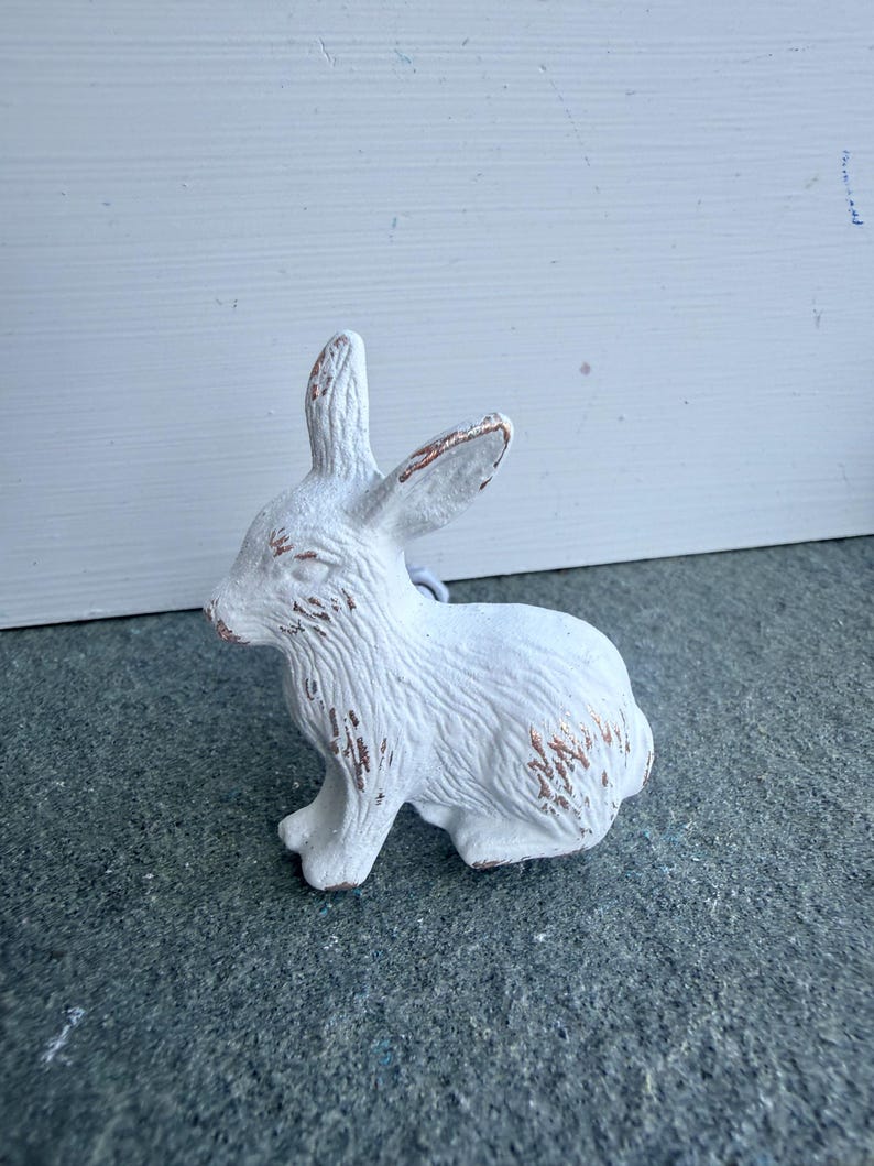 May include: A white rabbit figurine with a textured surface and visible brown distressed detailing on the ears, nose, and fur. The rabbit is depicted in a seated position, showing detailed fur patterns and pointed ears facing upward.