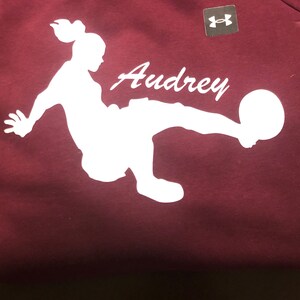 May include: Burgundy sweatshirt with a white silhouette of a person kicking a football. The name "Audrey" is written in white cursive above the silhouette. The Under Armour logo is in the top right corner.