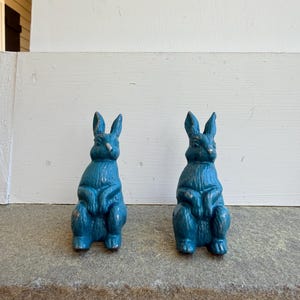 May include: Two teal-colored rabbit figurines with a distressed finish. The bunnies are in a seated position, with their ears upright. The figurines are placed on a gray surface, with a white painted wall in the background.