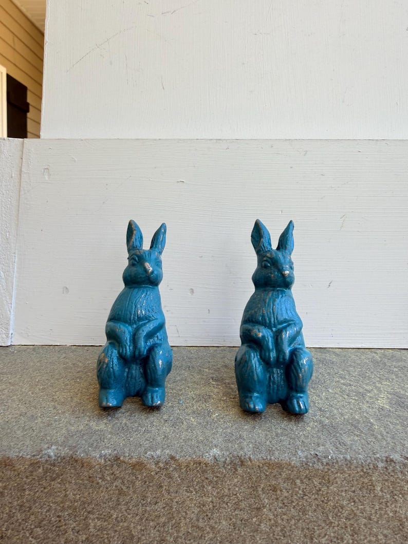 May include: Two teal-colored rabbit figurines with a distressed finish. The bunnies are in a seated position, with their ears upright. The figurines are placed on a gray surface, with a white painted wall in the background.