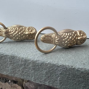 May include: Two gold-toned owl-shaped curtain tiebacks with rings. The owls have detailed features and are facing the same direction.