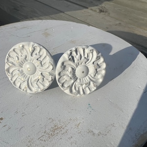May include: Two white, ornate, round, decorative knobs with a floral design. The knobs have a distressed, aged look and are made of metal.