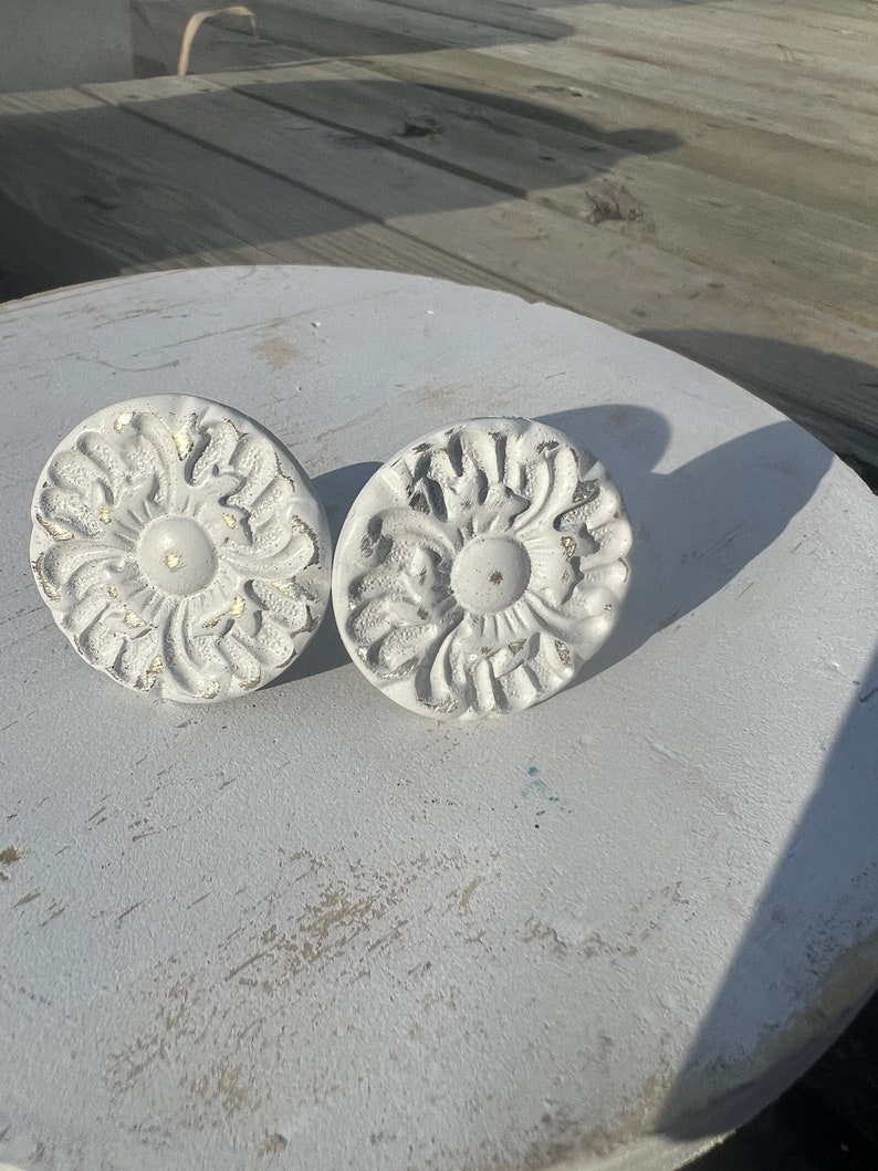 May include: Two white, ornate, round, decorative knobs with a floral design. The knobs have a distressed, aged look and are made of metal.