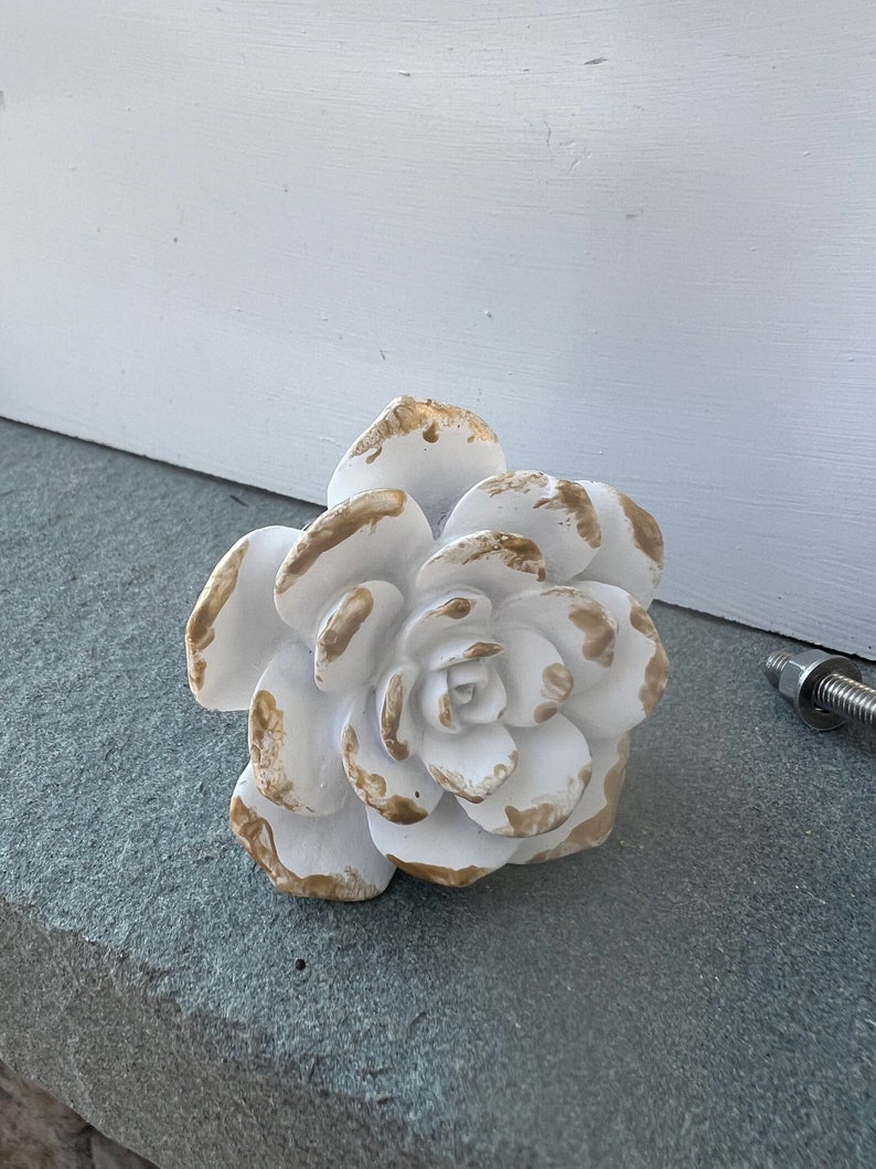 May include: White ceramic flower knob with gold accents. The flower has a detailed design and is perfect for adding a touch of elegance to any room.