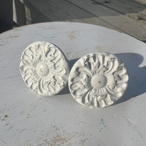 May include: Two white ceramic drawer pulls with a floral design. The pulls have a distressed white finish and a raised floral pattern.
