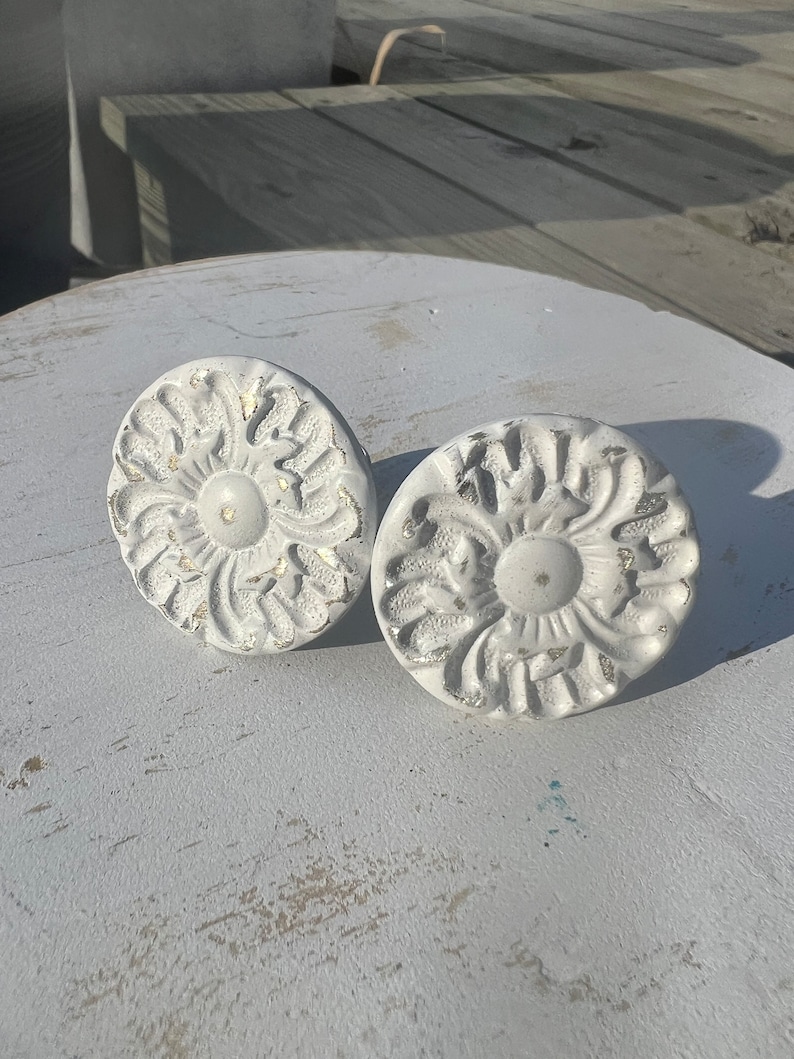 May include: Two white ceramic drawer pulls with a floral design. The pulls have a distressed white finish and a raised floral pattern.