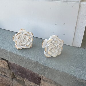 May include: Two white ceramic rose-shaped drawer pulls with gold accents. The pulls are on a grey stone surface.