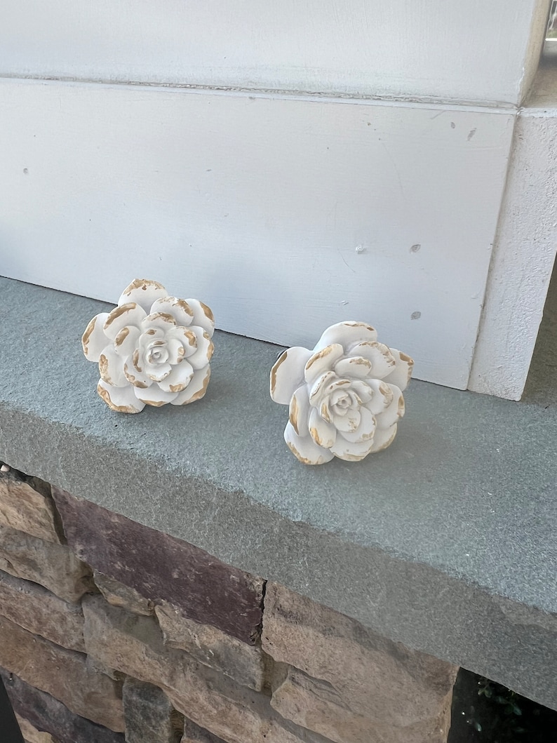 May include: Two white ceramic rose-shaped drawer pulls with gold accents. The pulls are on a grey stone surface.