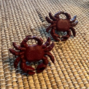 Op de afbeelding: Twee gietijzeren krabfiguren in een dieprode kleur. De krabben zijn gedetailleerd met scharen, poten en getextureerde schalen. Ze zijn geplaatst op een geweven, beige placemat, wat een kust- of nautisch thema cre&euml;ert.