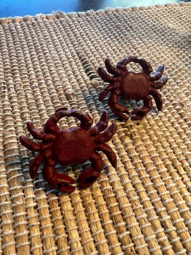 Op de afbeelding: Twee gietijzeren krabfiguren in een dieprode kleur. De krabben zijn gedetailleerd met scharen, poten en getextureerde schalen. Ze zijn geplaatst op een geweven, beige placemat, wat een kust- of nautisch thema cre&euml;ert.