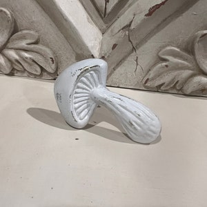 May include: White ceramic mushroom shaped hook. The hook is designed to look like a mushroom with a white stem and a white cap with ridges.