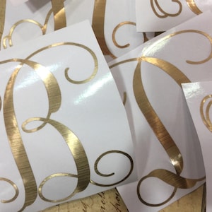 May include: Gold foil letter B with a decorative script font on a white background. The letter is a sticker or decal.