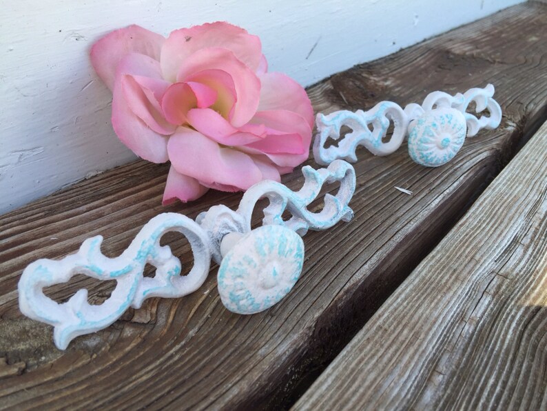 May include: Two white, ornate, decorative cabinet knobs with blue accents. The knobs have a floral design and are made of metal.
