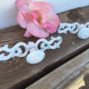 May include: Two white metal drawer pulls with ornate designs and blue painted accents. The pulls are on a wooden surface with a pink rose.