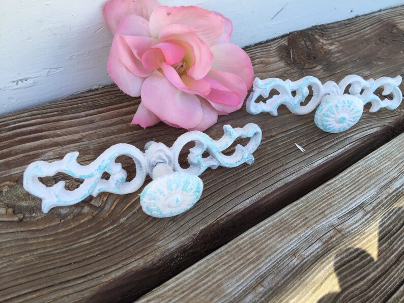 May include: Two white metal drawer pulls with ornate designs and blue painted accents. The pulls are on a wooden surface with a pink rose.