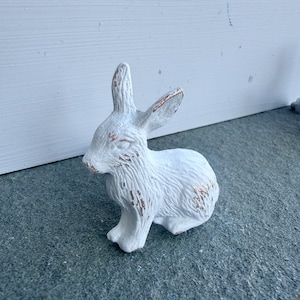 May include: A small, white, rabbit-shaped figurine with a textured surface and visible copper-colored distressing on the ears, face, and body. The rabbit is depicted in a seated position with its ears upright and alert, showcasing a weathered finish.