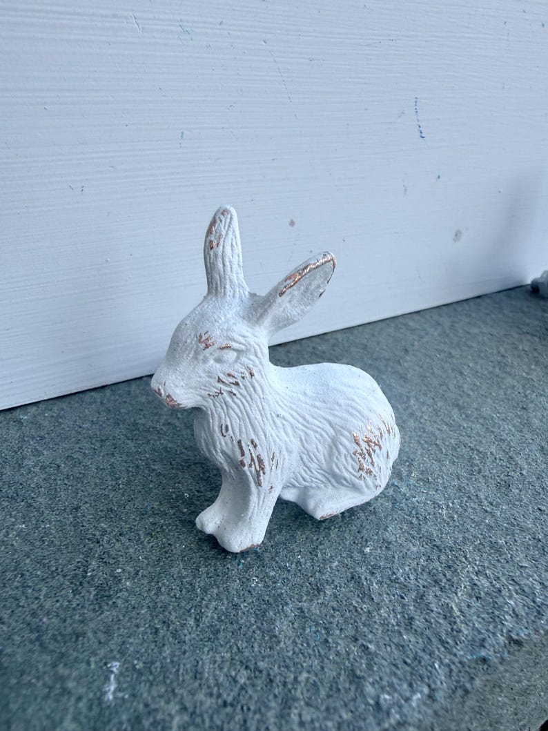 May include: A small, white, rabbit-shaped figurine with a textured surface and visible copper-colored distressing on the ears, face, and body. The rabbit is depicted in a seated position with its ears upright and alert, showcasing a weathered finish.