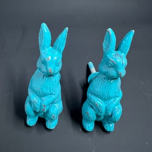 May include: Two turquoise blue ceramic rabbit figurines. The rabbits are sitting upright with their paws crossed. They have a distressed finish and are perfect for adding a touch of whimsy to your home decor.